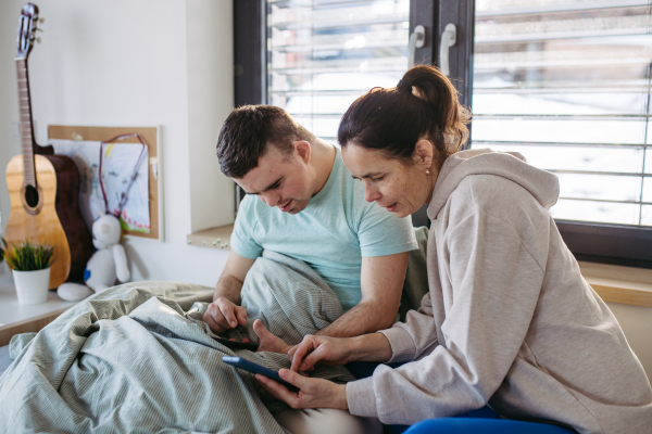 Young man with down syndrome sitting in his bed with mom, both looking at smartphone in the morning. Morning routine for man with disability.