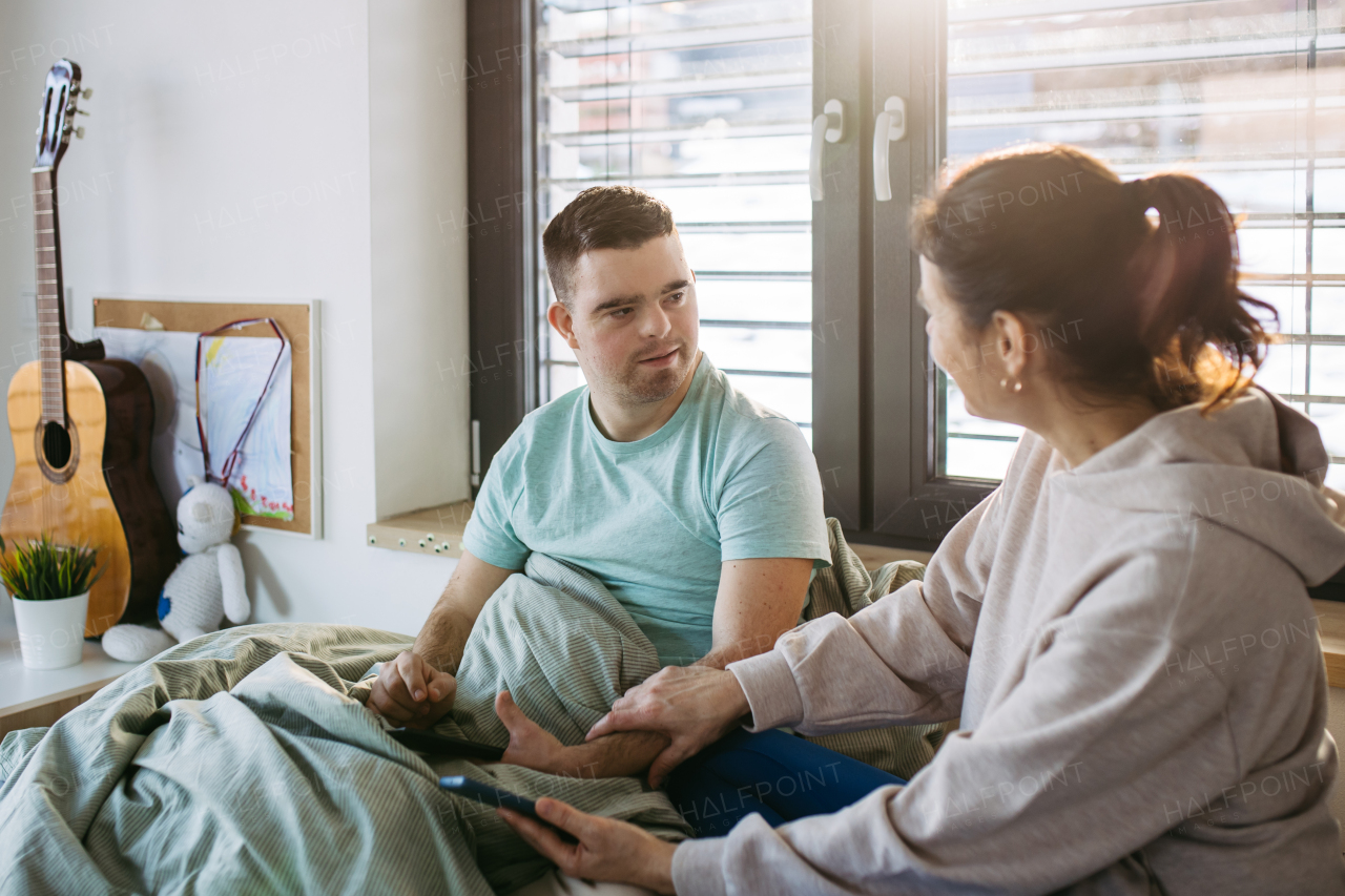 Young man with down syndrome sitting in his bed with mom, both looking at smartphone in the morning. Morning routine for man with disability.