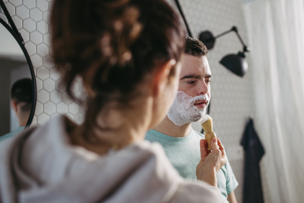 Mother applying shaving foam on son's face. Young man with down syndrome learning how to shave. Concept of mother's day and motherly love.