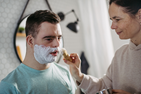 Mother applying shaving foam on son's face. Young man with down syndrome learning how to shave. Concept of mother's day and motherly love.