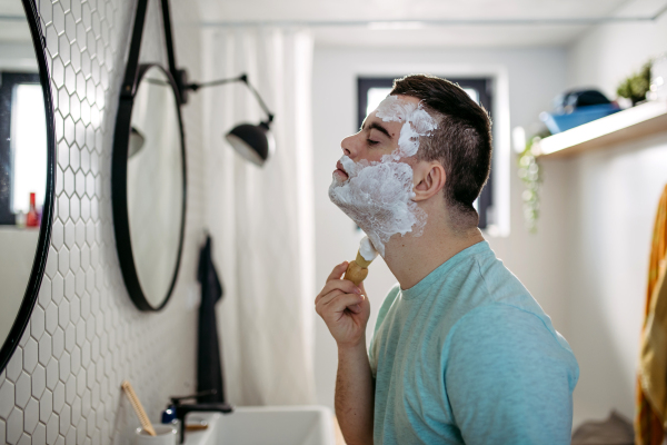 Young man with down syndrome learning how to shave, applying shaving foam, cream all over his face, looking at mirror.