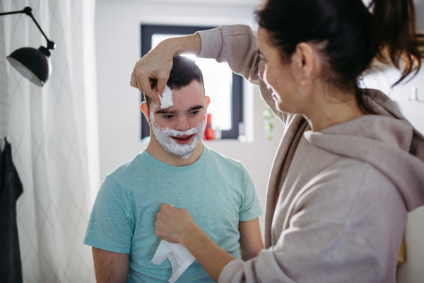 Mother applying shaving foam on son's face. Young man with down syndrome learning how to shave. Concept of mother's day and motherly love.