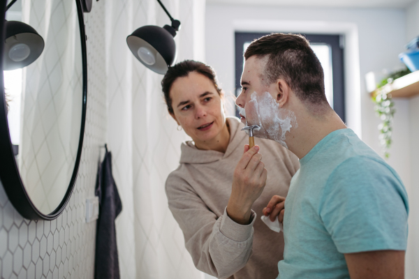 Mother applying shaving foam on son's face. Young man with down syndrome learning how to shave. Concept of mother's day and motherly love.