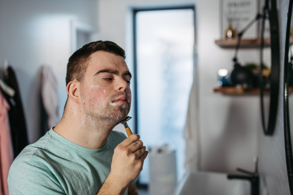 Young man with down syndrome learning how to shave, holding razor in hand, looking at mirror. Side view of focused disabled man.