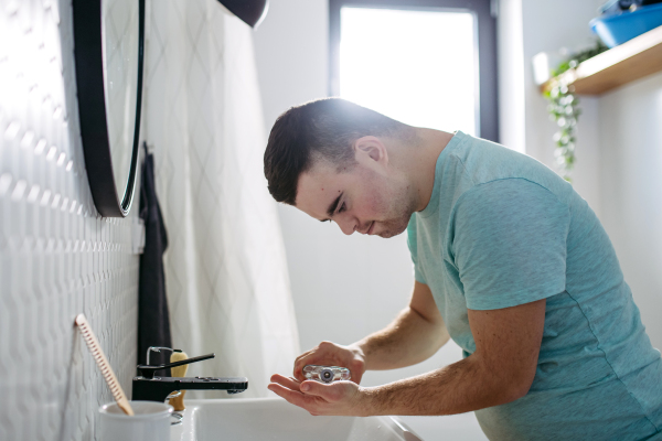 Young man with down syndrome learning how to shave,applying after shave lotion or cologne on face. Side view of focused disabled man.