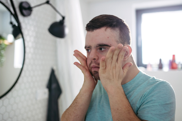 Rear view of young man with down syndrome in bathroom, holding his cheeks, going to shave his beard off.