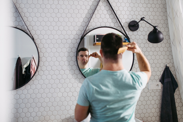 Rear view of young man with down syndrome in bathroom, combing his hairs.