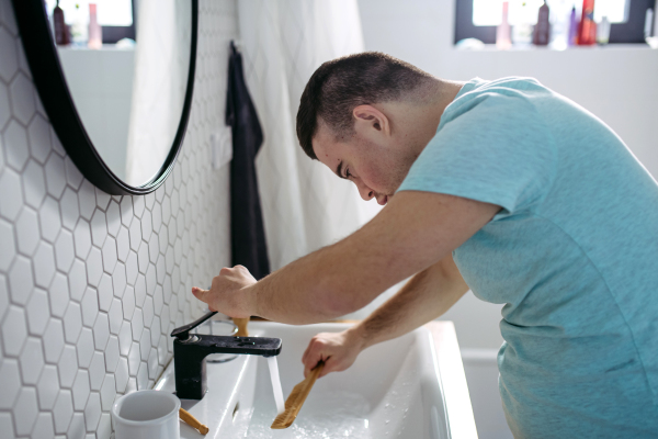 Side view of young man with down syndrome in bathroom, cleaning comb, rinsing it in the water.