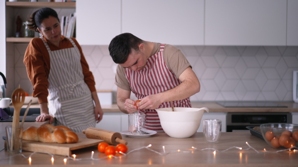 Young man with Down syndrome preparing breakfast with his mother at home, cracking eggs. Morning routine for man with Down syndrome genetic disorder.