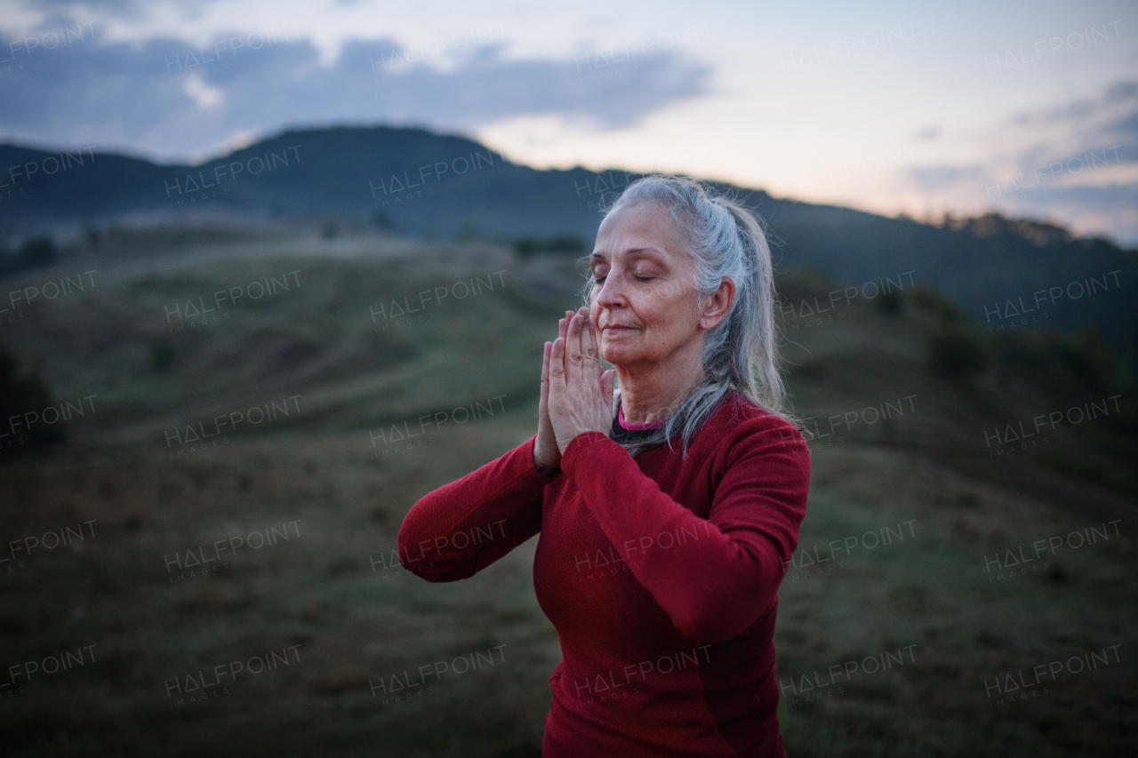 A senior woman doing breathing exercise in nature on early morning with fog and mountains in background.