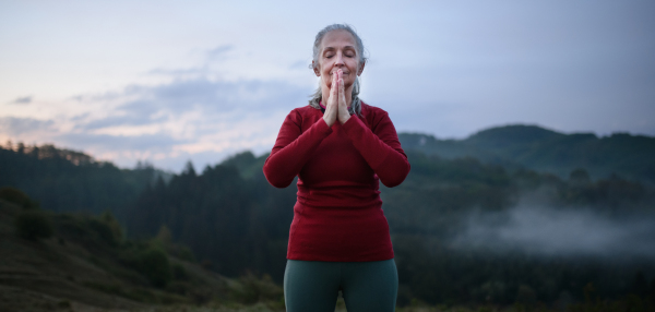 A senior woman doing breathing exercise in nature on early morning with fog and mountains in background.