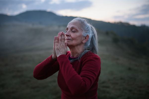 A senior woman doing breathing exercise in nature on early morning with fog and mountains in background.