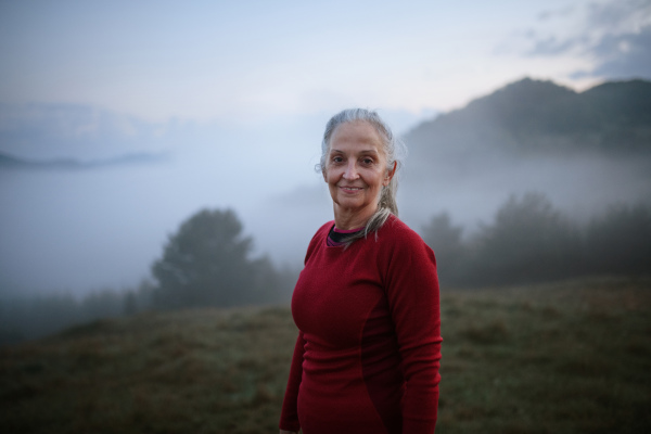 A cheerful senior woman in nature on early morning with fog and mountains in background.