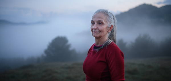 A cheerful senior woman in nature on early morning with fog and mountains in background.