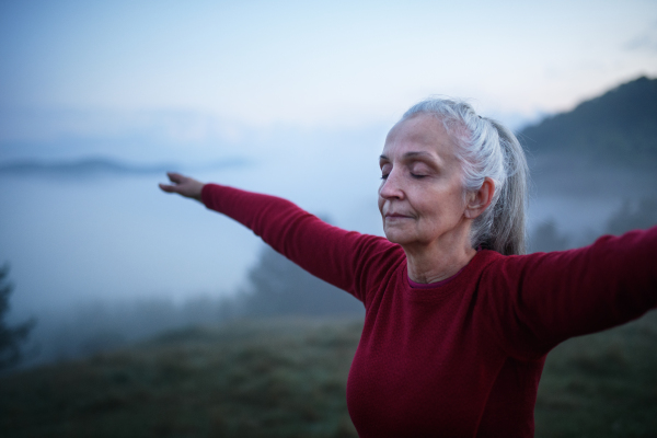 A senior woman doing breathing exercise in nature on early morning with fog and mountains in background.