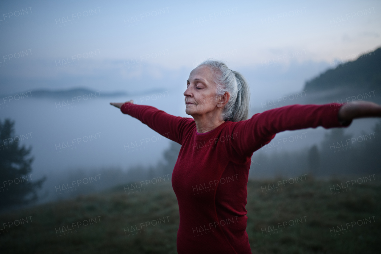 A senior woman doing breathing exercise in nature on early morning with fog and mountains in background.