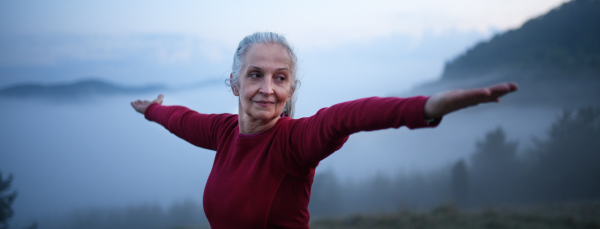 A senior woman doing breathing exercise in nature on early morning with fog and mountains in background.