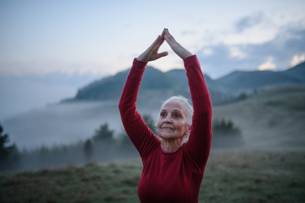 A senior woman doing breathing exercise in nature on early morning with fog and mountains in background.