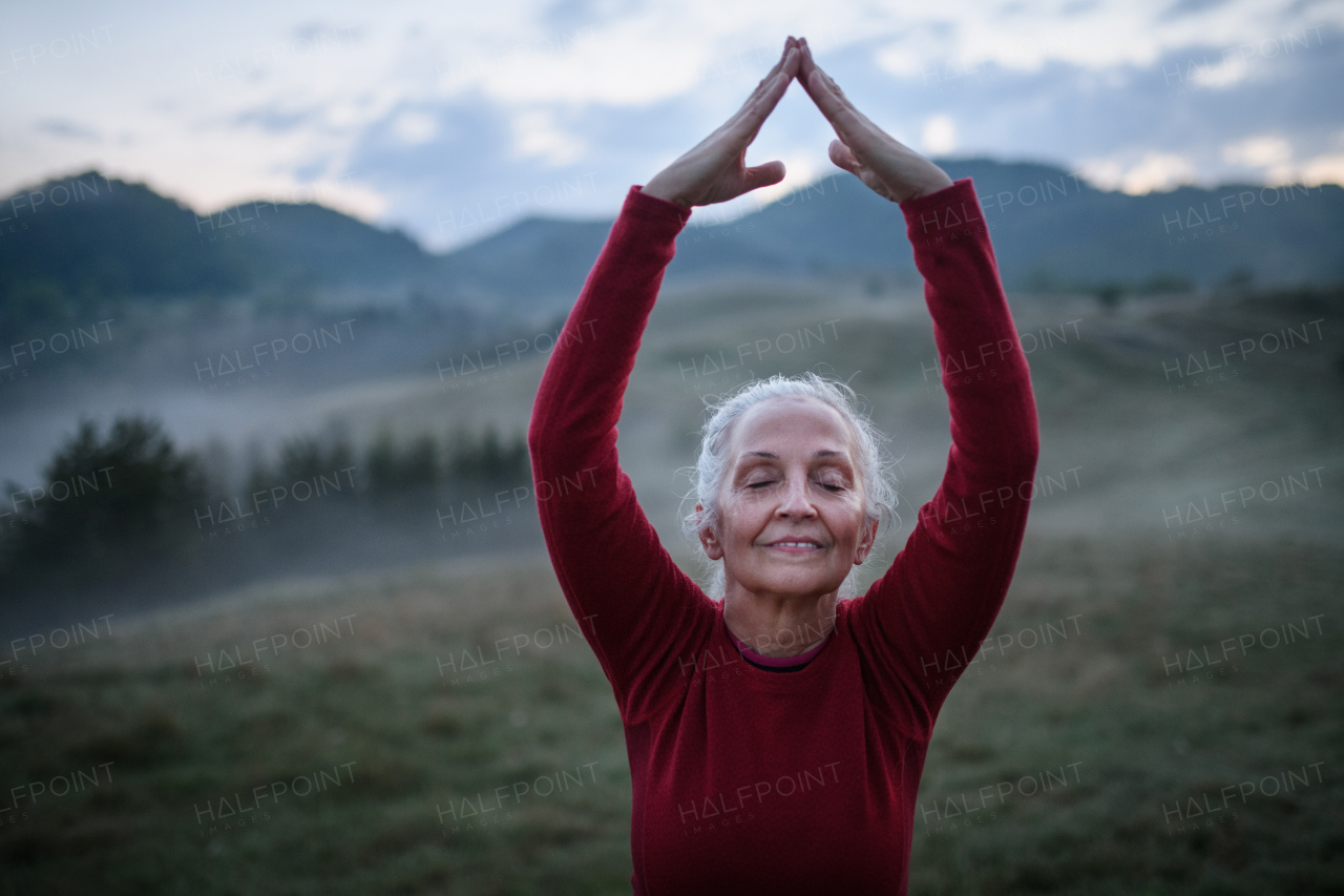 A senior woman doing breathing exercise in nature on early morning with fog and mountains in background.