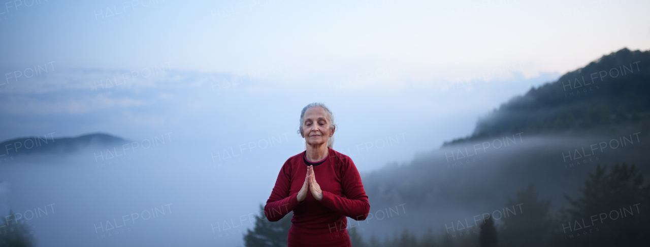 A senior woman doing breathing exercise in nature on early morning with fog and mountains in background.