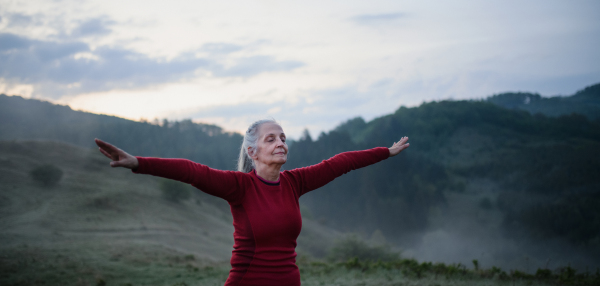 A senior woman doing breathing exercise in nature on early morning with fog and mountains in background.