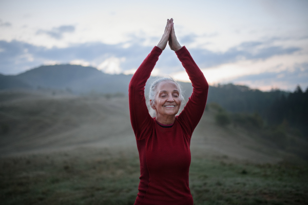 A senior woman doing breathing exercise in nature on early morning with fog and mountains in background.