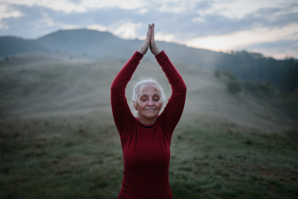 A senior woman doing breathing exercise in nature on early morning with fog and mountains in background.