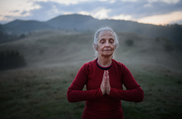 A senior woman doing breathing exercise in nature on early morning with fog and mountains in background.