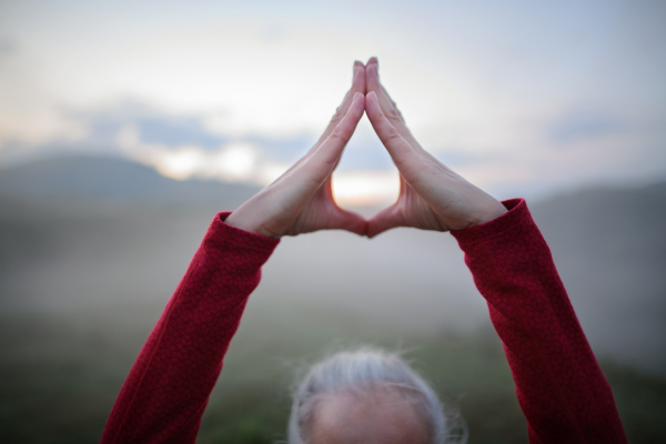 A senior woman doing breathing exercise in nature on early morning with fog and mountains in background.