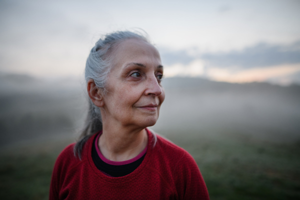 A cheerful senior woman in nature on early morning with fog and mountains in background.