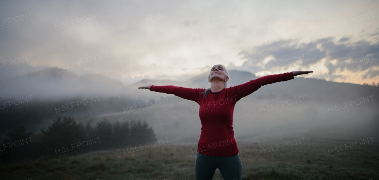 A senior woman doing breathing exercise in nature on early morning with fog and mountains in background.