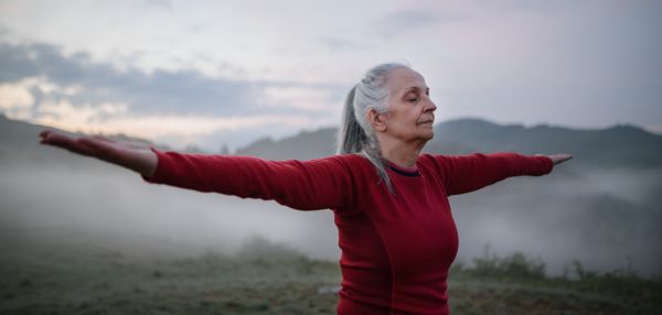 A senior woman doing breathing exercise in nature on early morning with fog and mountains in background.