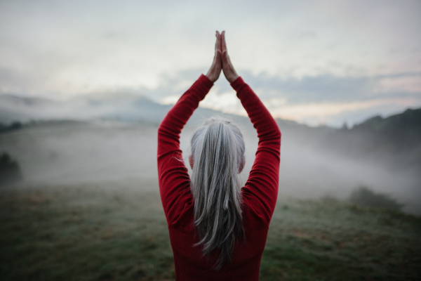 A rear view of senior woman doing breathing exercise in nature on early morning with fog and mountains in background.