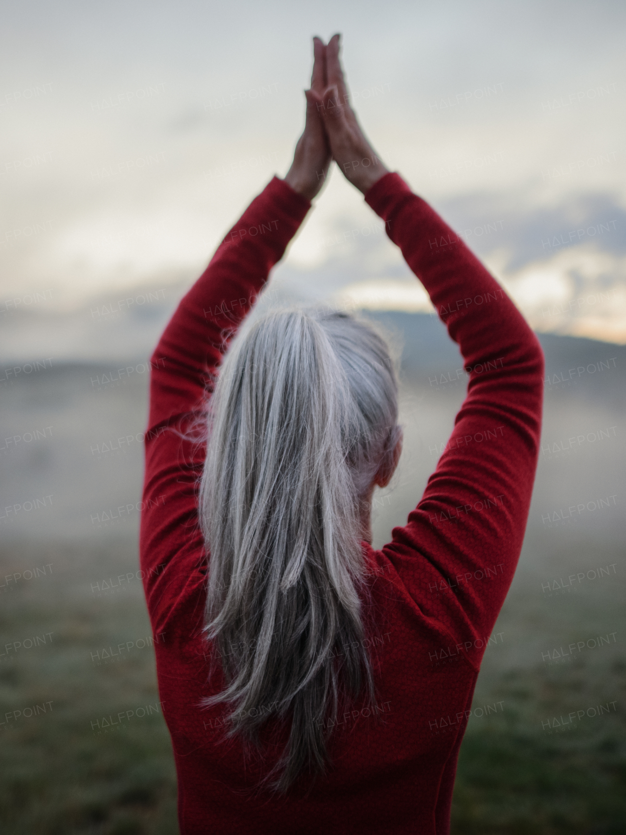 A rear view of senior woman doing breathing exercise in nature on early morning with fog and mountains in background.