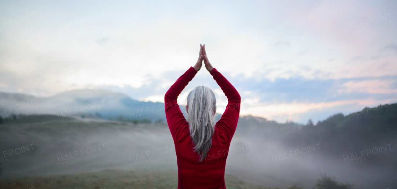 A rear view of senior woman doing breathing exercise in nature on early morning with fog and mountains in background.