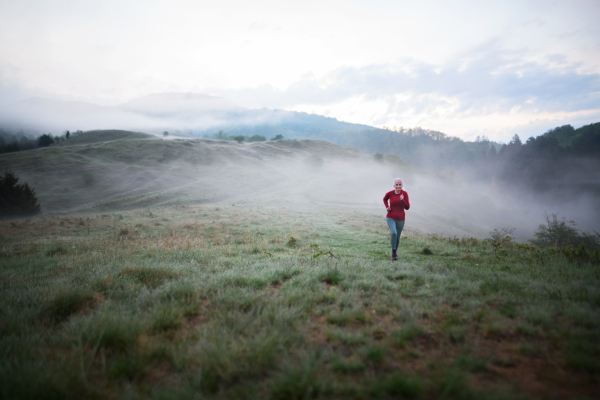 A senior woman jogging in nature on early morning with fog and mountains in background.