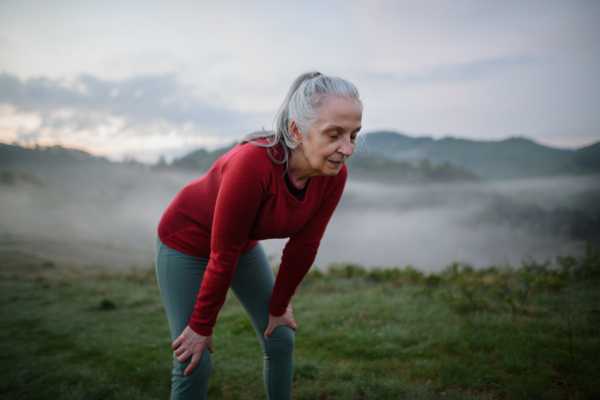 A senior woman doing breathing exercise in nature on early morning with fog and mountains in background.