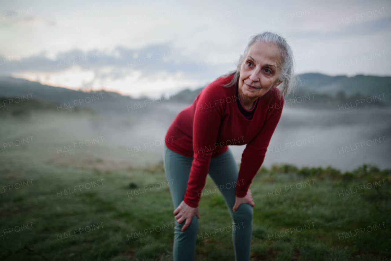 A senior woman doing breathing exercise in nature on early morning with fog and mountains in background.