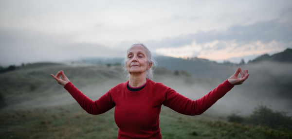 A senior woman doing breathing exercise in nature on early morning with fog and mountains in background.