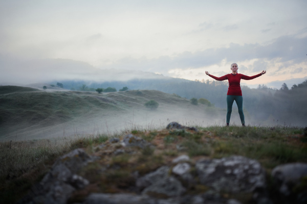 A senior woman doing breathing exercise in nature on early morning with fog and mountains in background.