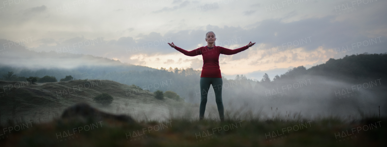 A senior woman doing breathing exercise in nature on early morning with fog and mountains in background.