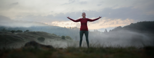 A senior woman doing breathing exercise in nature on early morning with fog and mountains in background.