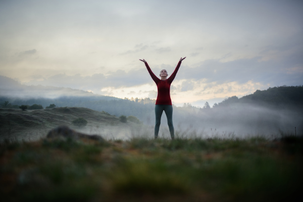 A senior woman doing breathing exercise in nature on early morning with fog and mountains in background.