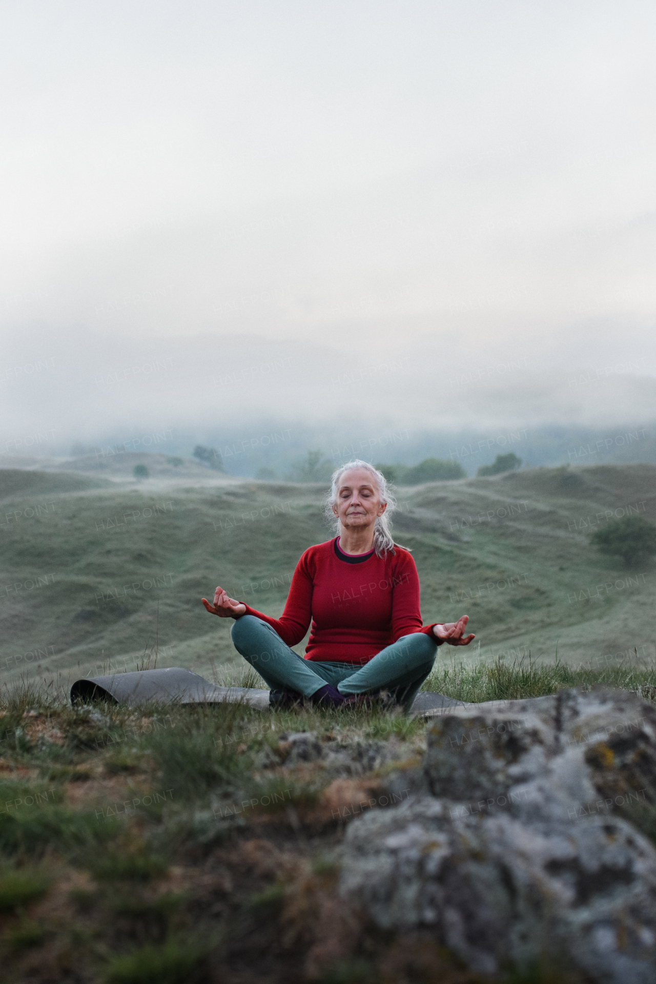 A senior woman doing breathing exercise in nature on early morning with fog and mountains in background.