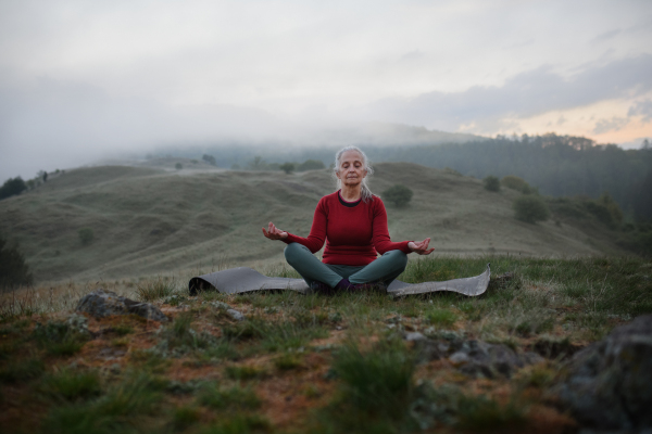 A senior woman doing breathing exercise in nature on early morning with fog and mountains in background.