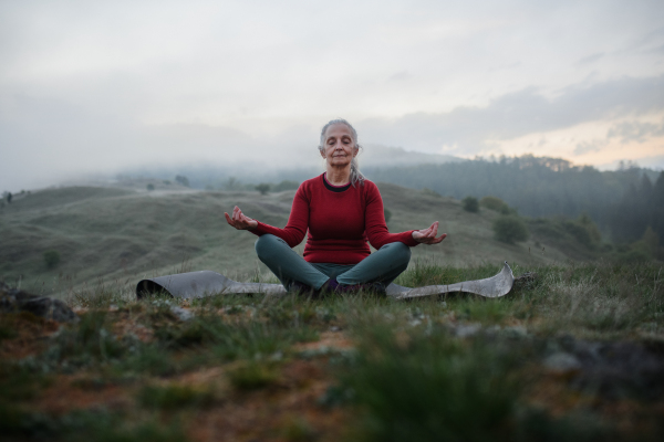 A senior woman doing breathing exercise in nature on early morning with fog and mountains in background.