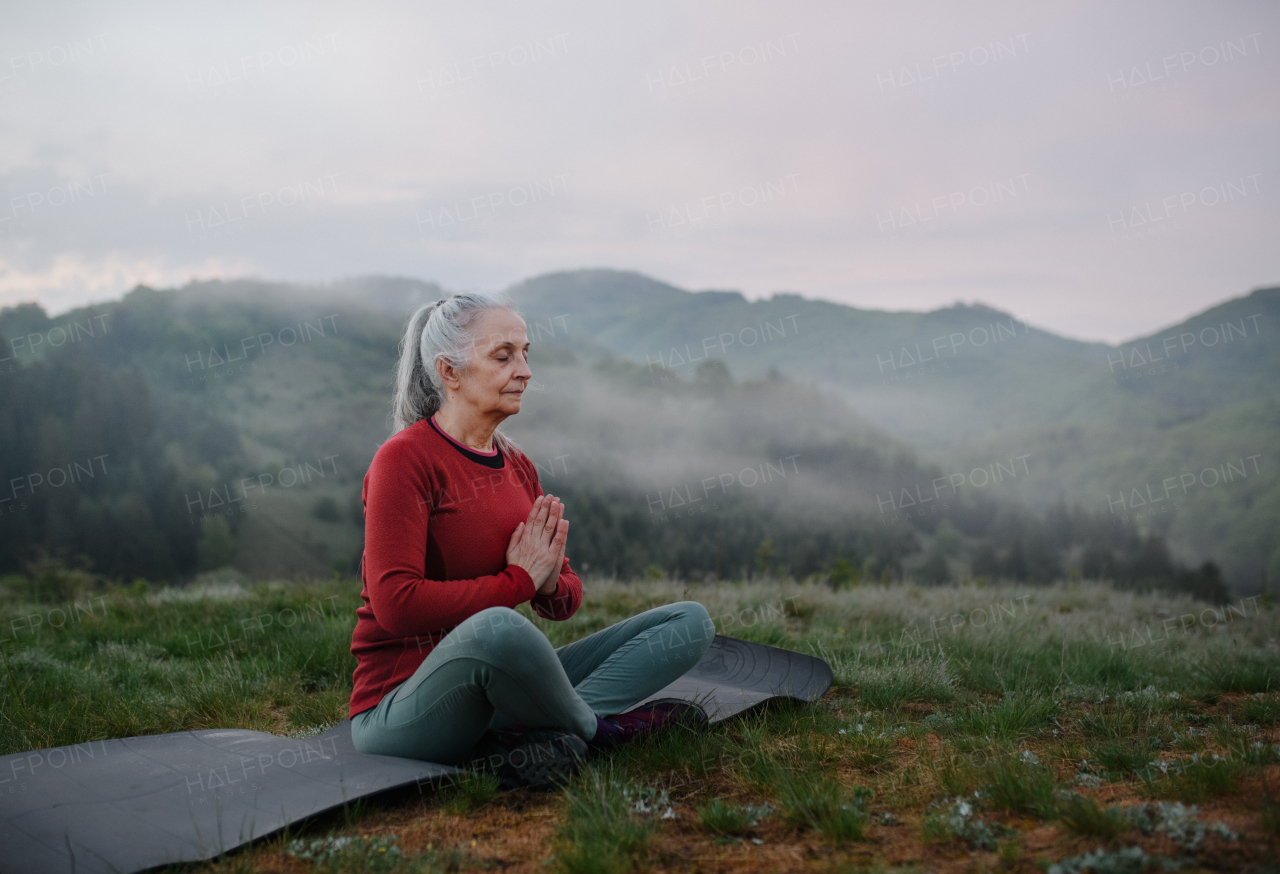 A senior woman doing breathing exercise in nature on early morning with fog and mountains in background.