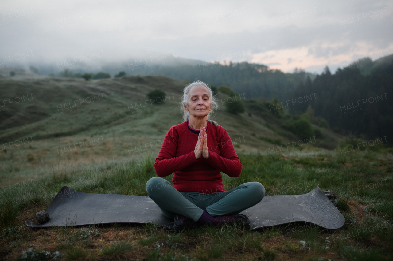 A senior woman doing breathing exercise in nature on early morning with fog and mountains in background.