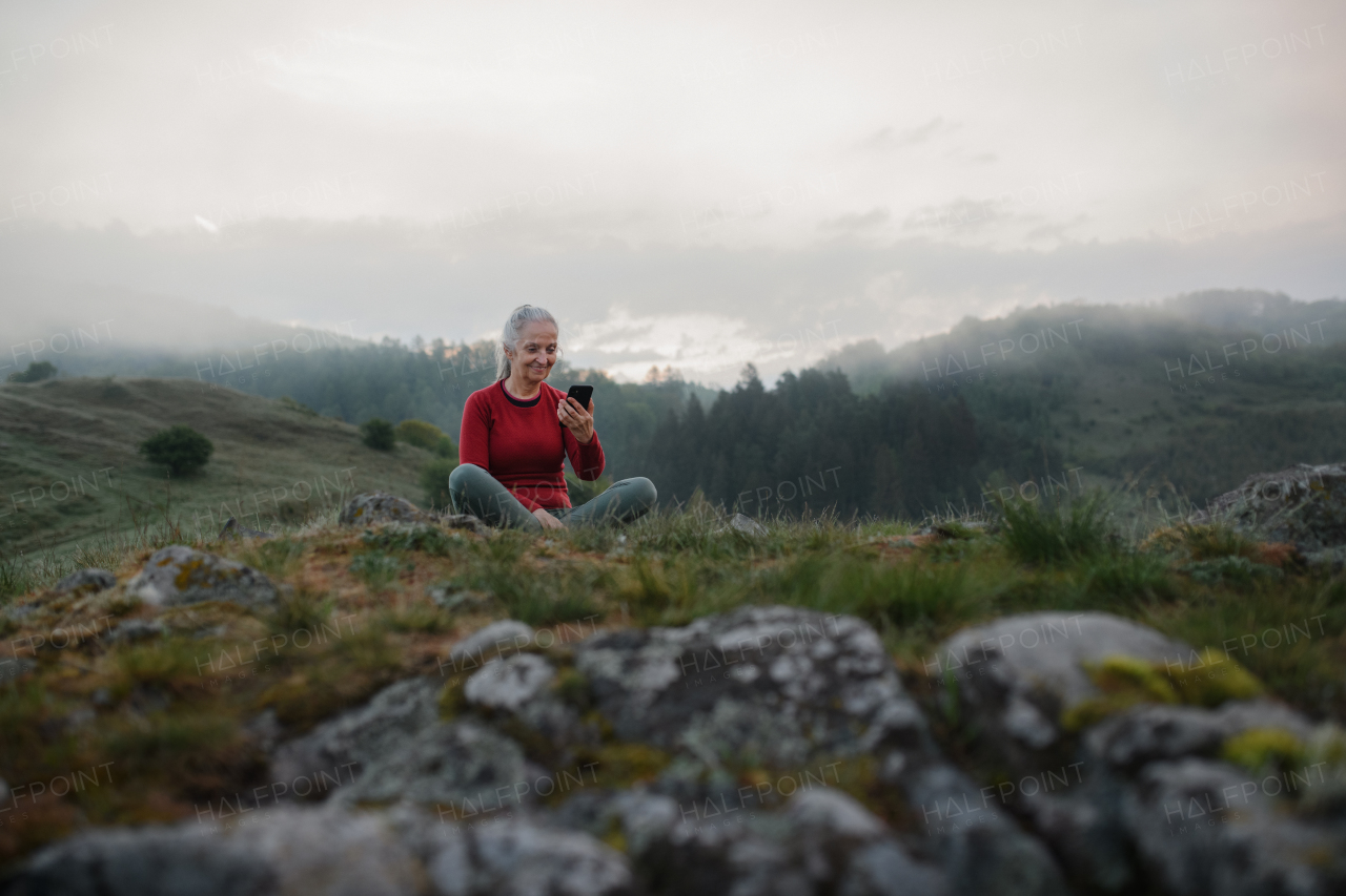 A senior woman taking selfie when doing breathing exercise in nature on early morning with fog and mountains in background.