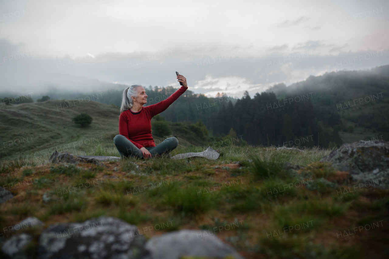 A senior woman taking selfie when doing breathing exercise in nature on early morning with fog and mountains in background.